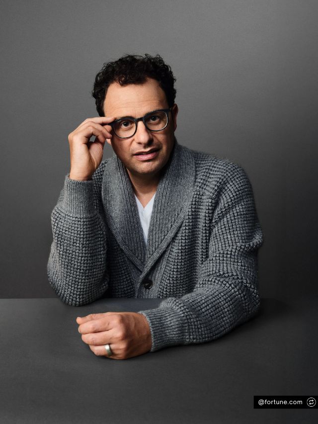 Anthropic Co-Founder Dario Amodei Net Worth, Journey, Vision and Growth 2 Dario Amodei, co-founder of Anthropic, adjusts his glasses while wearing a cozy grey cardigan and posing against a dark backdrop.