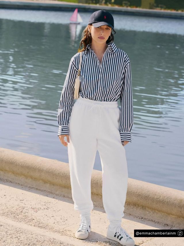 Chamberlain Coffee Founder Emma Chamberlain Net Worth, Journey, Vision & Growth 2 Emma Chamberlain, founder of Chamberlain Coffee, poses by a serene lake wearing white joggers and a classic striped button-up.