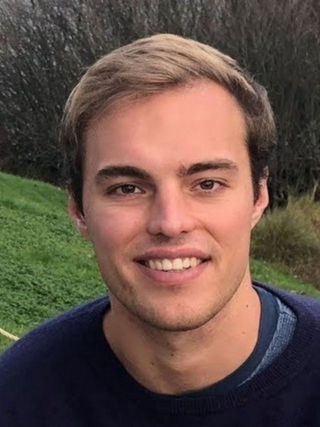 Nourish Co-Founder Aidan Dewar, Journey in Digital Health & Nutrition 2 Aidan Dewar, co-founder of Nourish, smiles brightly outdoors while wearing a casual dark blue sweater over a crewneck tee.